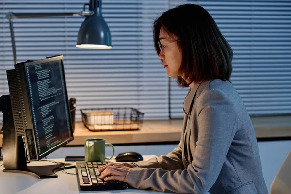 Asian IT specialist typing codes for program on computer working at her workplace till late evening