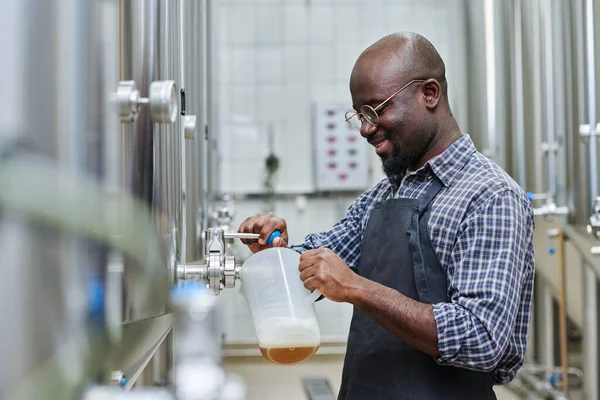 stock image African American factory worker pouring brewed beer in container for tasting after processing