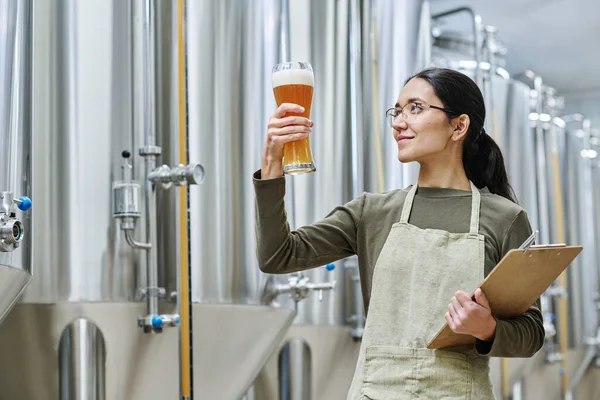 Specialist with document looking at glass of fresh beer to check the quality during her work at brewing plant