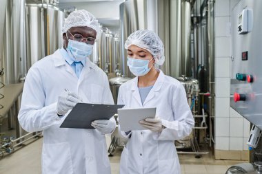 Maskeli ve beyaz önlüklü mühendisler tablet bilgisayar üzerinde çalışıyor ve fabrikadaki işleri sırasında belgelere not alıyorlar.