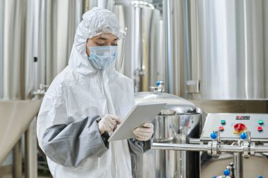 Engineer in protective wear working online on digital tablet at chemical factory clipart