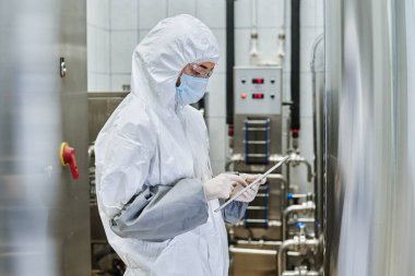 Fabrikanın atölyesinde duran tablet bilgisayardaki işi kontrol eden koruyucu kostümlü bir mühendis.