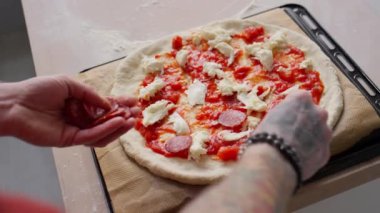 Erkek şefin omuzlarının üzerinden domates soslu pizzanın üstüne parçalanmış mozzarella peyniri ve kağıt kaplı biberli pepperoni koyarken.