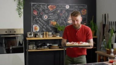 Sakallı erkek yemek blogcusu kameraya bakıyor, konuşuyor ve sonra fırına pizza katıyor ve mutfakta online pişirme dersi veriyor.