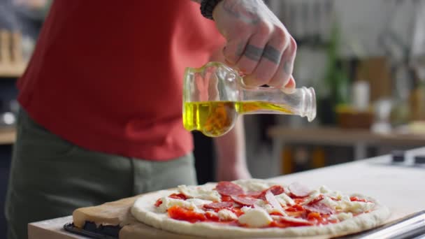 キッチンで調理中に男性食品ブロガーの撮影は ベーキング前にピザの上にオリーブオイルを注ぎ カメラで悪魔ホーン手ジェスチャーを示します — ストック動画