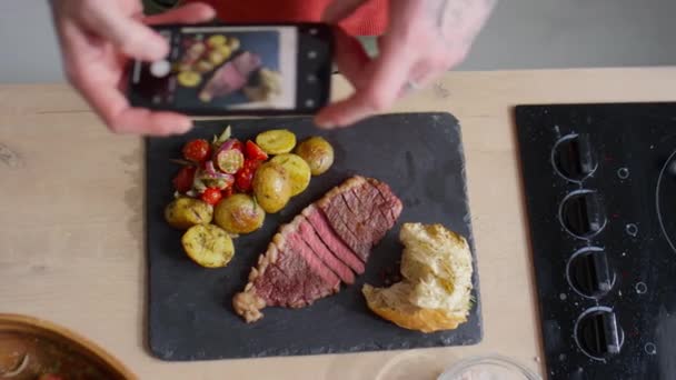 スマートフォンで男性フードブロガーの手のトップダウンショットを撮影しながら スレートボード上で提供されるジャガイモ 野菜サラダやパンと牛肉のステーキの写真を撮る — ストック動画