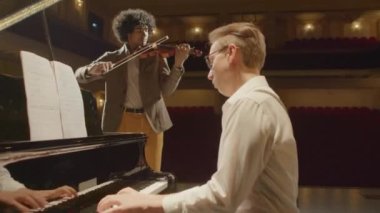 Kafkasyalı müzisyen konser salonundaki prova sırasında sahnede keman çalan Afrikalı Amerikalı adam olarak piyano çalıyor.