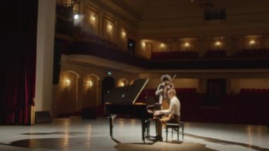 İki profesyonel müzisyenin konser salonunda hiç seyirci olmadan keman ve kuyruklu piyano çaldığı geniş açılı bir çekim.