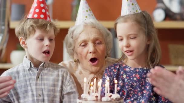 Oudere Vrouw Met Feestmuts Omarmt Kleinkinderen Blaast Kaarsen Verjaardagstaart Samen — Stockvideo