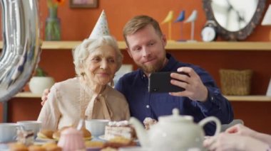 Neşeli Kafkasyalı adam parti şapkalı neşeli büyükanneyi kucaklıyor, evde doğum günü yemeğinde akıllı telefonuyla selfie çekiyor ve gülümsüyor.