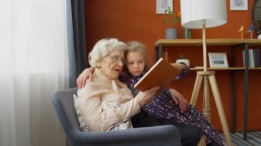 Yaşlı büyükannenin küçük torunuyla koltukta oturduğu ve evde birlikte kitap okuduğu fotoğrafı yakınlaştır.