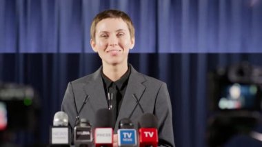 Kadın konuşmacı, basın toplantısı sırasında poz verirken mikrofonla podyumda mutlu bir şekilde gülümsüyor.