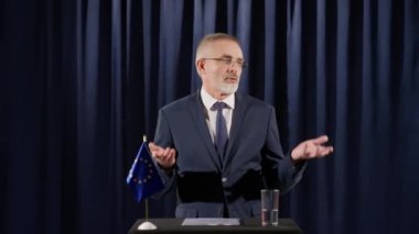 Üst düzey erkek siyasetçi AB bayrağıyla kürsüde duruyor ve basın toplantısında gazetecilere sesleniyor