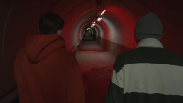 赤い光を持つ暗いトンネルを一緒に歩くストリートウェアで2人の友人のショットの後 — ストック動画