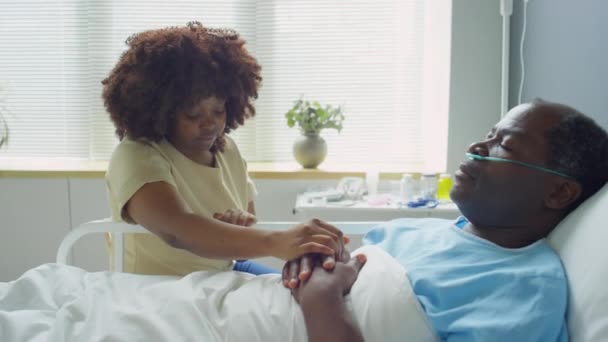 放大镜头拍摄的有关非洲裔美国人妻子坐在医院病房的床边 在接受治疗时牵着不省人事的生病丈夫的手 — 图库视频影像
