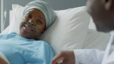 Burun kanülü ve kafa sargılı genç Afro-Amerikan kadın hastaların odak noktası. Doktoru dinliyor ve hastane yatağında yatarken gülümsüyor.
