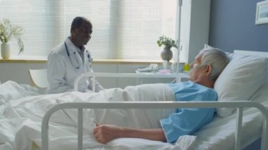 Afro-Amerikan doktorların hastane koğuşunda yatarken burun kanülü olan yaşlı beyaz bir hastayla konuşurken görüntüsünü yakınlaştırın.