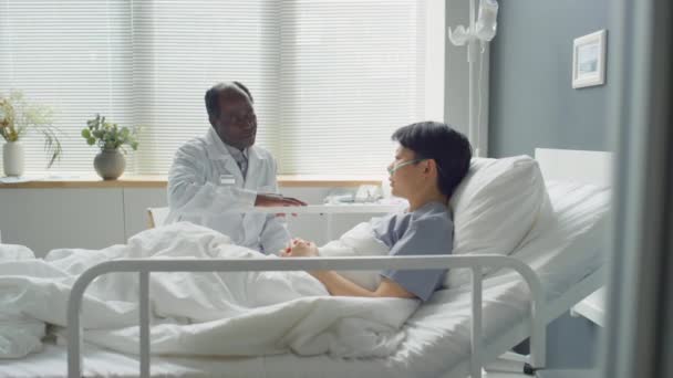 放大非洲裔美国医生坐在医院病房病床上的镜头 与亚洲女性鼻腔炎患者讨论治疗方案 — 图库视频影像