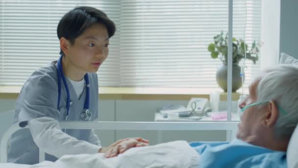 若いアジアの女性医師は 治療中に彼をサポートしながら 病院のベッドの上に横に高齢者の白人患者の話すと手を打つ — ストック動画