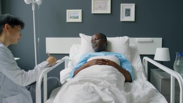 放大拍摄的非洲裔美国病人躺在医院病房的床上 在她做笔记的时候与亚洲女医生交谈 — 图库视频影像