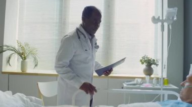 Afro-Amerikan doktorların hastane koğuşunda yatarken burun kanülü olan yaşlı beyaz bir hastayla konuşurken görüntüsünü büyüt.