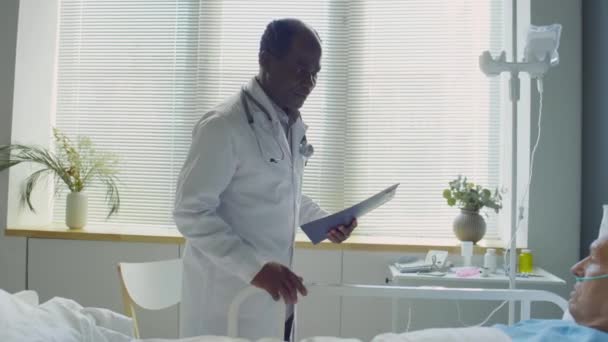 アフリカ系アメリカ人の医者が病院の病棟を歩いているところをズームアウトして ベッドに座って 鼻カニューレと横たわっている上級白人患者と話をする — ストック動画