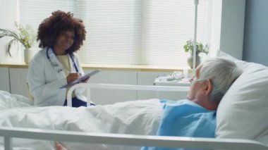 Laboratuvar önlüklü genç Afrikalı kadın doktor yaşlı beyaz bir hastayla konuşuyor, gülümsüyor ve hastane koğuşunda onu kontrol ederken not alıyor.