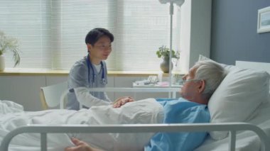 Genç Asyalı kadın doktor yaşlı beyaz bir hastanın elini okşuyor, hastanede tedavi sırasında gülümsüyor ve onunla konuşuyor.