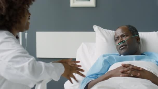 病院の病棟でベッドに座って治療中に彼をサポートしながら男性患者とチャットしている陽気なアフリカ系アメリカ人女性医師 — ストック動画