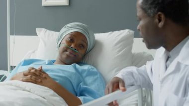 Burun kanülü ve kafa bandı olan Afro-Amerikalı kadın hastane yatağında uzanıyor ve ziyaretinde doktorla konuşuyor.