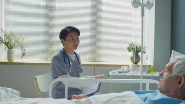 放大拍摄身穿医疗制服的年轻亚洲女医生在医院病房里行走 坐在床边 与白种人老人交谈并做笔记的镜头 — 图库视频影像