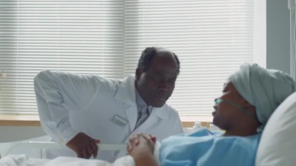 アフリカ系アメリカ人の医師が病院の病棟を歩き ベッドサイドに座り 若い女性患者とヘッドラップで話す — ストック動画