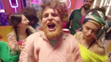 Genç, çakırkeyif, sakallı, kızlarla dans eden ve parıldayan neon süslemelerle dolu bir odada kamera önünde şarkı söyleyen genç bir adam.