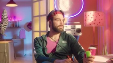 Yeşil spor ceketli, pembe kolsuz tişörtlü, spor bantlı, elinde tenis topu tutan ve neon dekorasyonlu bir stüdyoda kameraya poz veren genç sakallı adamın portre fotoğrafını yakınlaştır.