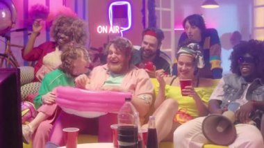 Çok ırklı giyinmiş, içki içen, abur cubur yiyen ve TV 'de neon ışıklı komedi filmi seyreden çok yönlü arkadaşlara yakınlaşın.