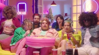 Moda kıyafetleriyle kanepeye oturmuş, renkli neon ışıklarıyla parti odasında korku filmi izlerken kameraya bakıp korkan çok ırklı genç arkadaşları yakınlaştırın.