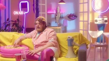 Kanepede oturan parlak kıyafetli genç adamı, renkli neon süslemeleri olan odada televizyon izlerken patlamış mısır yerken ve gülümserken çek.
