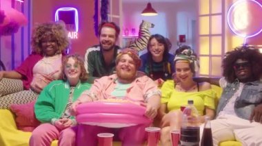 Moda kıyafetleri içinde, patlamış mısır ve içeceklerle kanepede oturmuş neon ışıklı komedi filmi izlerken kameraya bakıp gülen neşeli arkadaşlara yakınlaşın.