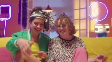 Moda kıyafetleri içinde, kanepede oturmuş patlamış mısır yiyen, sohbet eden ve neon süslemeli televizyon seyreden iki neşeli kız arkadaşın görüntüsünü yakınlaştırın.