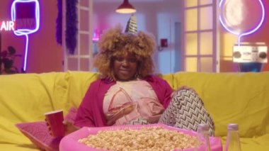 Afro-Amerikan bir kızın parlak kıyafetiyle kanepeye uzanırken, içki ve patlamış mısırla, kameraya bakarken ve neon süslemelerle parti odasında poz verirken görüntüsünü büyüt.