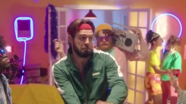Yeşil eşofman ve fitness saç bandı takmış genç sakallı adamın 90 'ların disko partisinde neon ışıklı odada arkadaşlarıyla dans ederken görüntüsünü yakınlaştır.