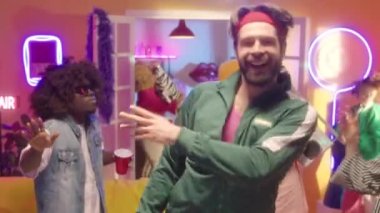 Yeşil eşofman ve fitness saç bandı giyen mutlu sakallı adamın görüntüsünü yakınlaştır ve 90 'ların disko partisinde neon ışıklı arkadaşlarıyla enerjik bir şekilde dans et.