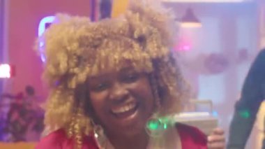 Parlak fuşya kıyafetli mutlu Afrikalı kız elinde plastik bardakla, gülümseyip dans ederken arkadaşlarıyla disko partisinde eğleniyor.