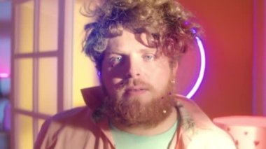 Disko ışıkları ve neon süslemelerle dans ederken kameraya gülümseyen parlak elbiseli, sakallı neşeli adamın portresi