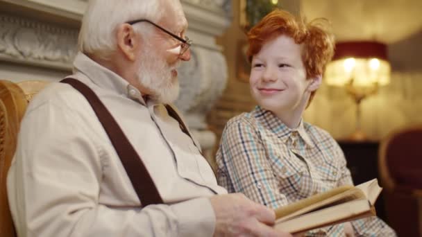 Старший Дід Сидячи Щасливим Онуком Біля Каміна Вдома Читаючи Книгу — стокове відео