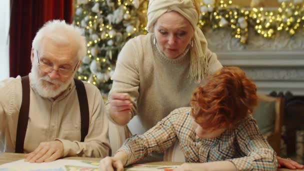 小男孩和爷爷用蜡笔在纸上画画 就像快乐的奶奶在用圣诞树和神灯装饰的客厅里告诉他们一些事情一样 — 图库视频影像