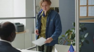 Beyaz bir genç, Amerikan elçiliğinde yürüyor, Afrikalı Amerikan konsolosluğuna belgeler veriyor ve vize görüşmesi sırasında sorularını cevaplıyor.