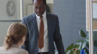 Resmi takım elbiseli Afro-Amerikalı adam AB elçiliğinde yürüyor, kadın memurlara belgeler veriyor ve Shengen vizesine başvururken röportaj yapıyor