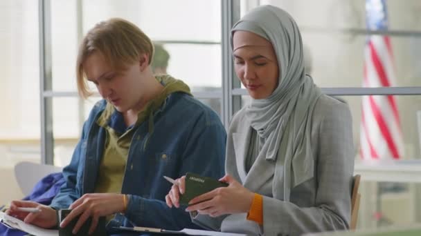 头戴头巾的穆斯林妇女和年轻的白人男子一起坐在美国大使馆填写签证申请表 — 图库视频影像