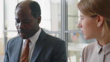 Resmi giyinen Afrikalı Amerikalı işadamı, yabancı elçilikteki vize görüşmesinden önce sırada beklerken beyaz bir kadınla konuşuyor.